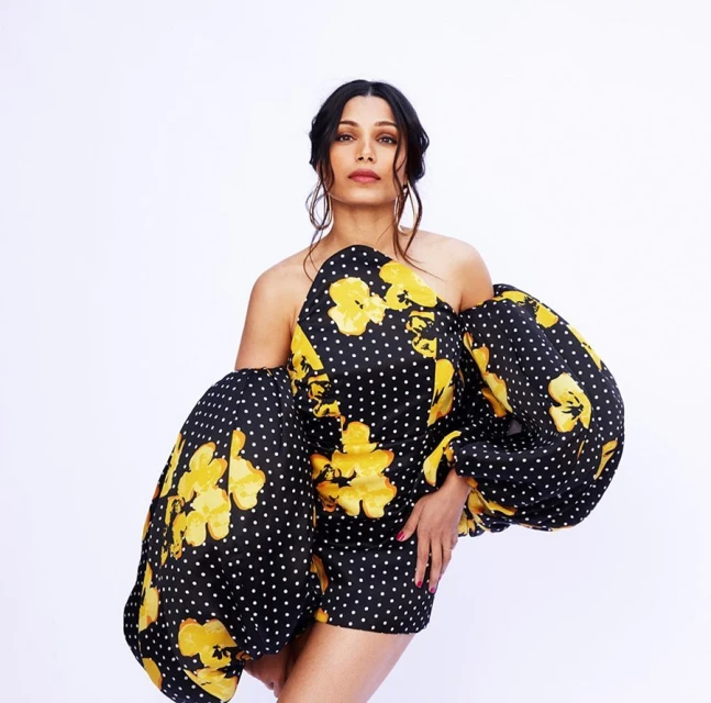Portrait of Freida Pinto wearing a black and yellow floral off-the-shoulder dress against a white background in a studio shot.