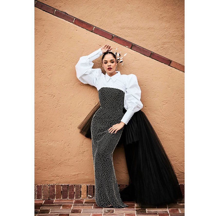 Elegant woman in embellished gown and white shirt posing against a tan wall with a fashionable headpiece for a Beauty editorial.
