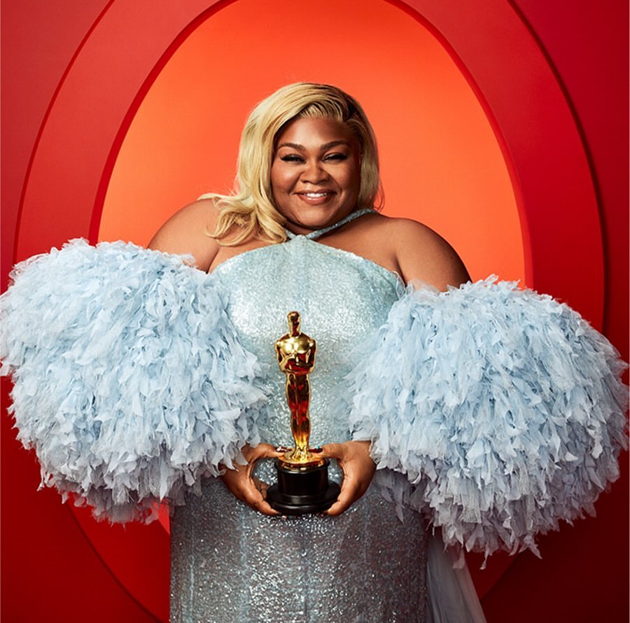 Smiling, plus-size adult female with blond hair holds a gold Oscar statue. She wears a light blue dress with feathered sleeves.