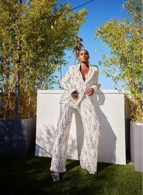 Adult woman in a sequined pantsuit poses outdoors with planters and sky background, creating a fashion editorial shot in natural light.