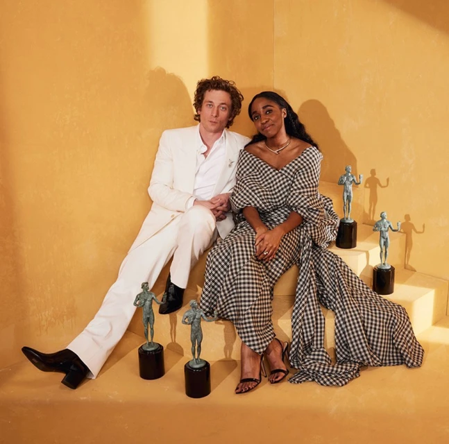 A man in a white suit and a woman in a black and white dress sitting on stairs with bronze statues in a bright, golden-hued room.