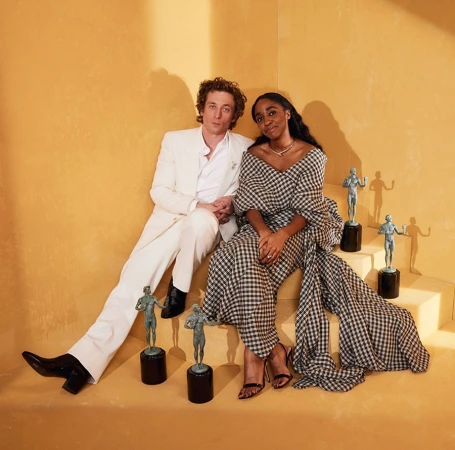 A man in a white suit and a woman in a black and white dress sitting on stairs with bronze statues in a bright, golden-hued room.