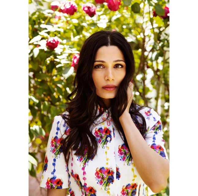Portrait of Freida Pinto against a backdrop of red roses, wearing a floral patterned top and touching her hair in natural light.