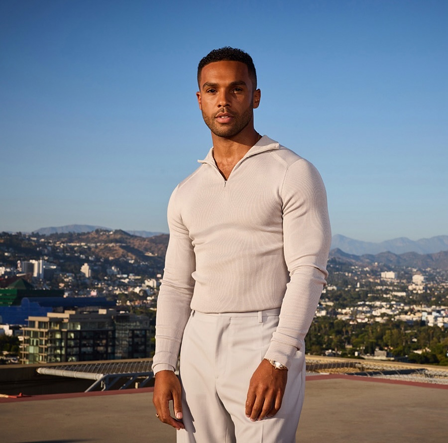 Portrait of a Black male adult wearing a ribbed shirt and trousers on a rooftop with Los Angeles cityscape in the background.
