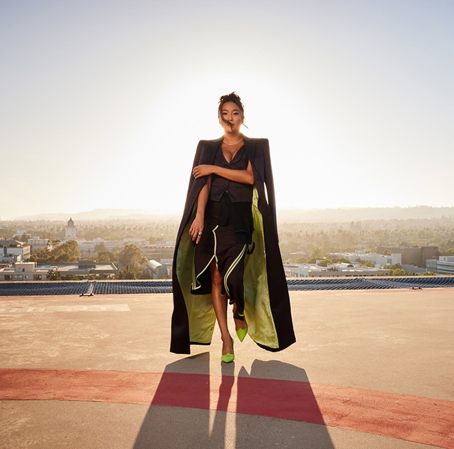Elegant woman in a black coat and skirt, accented by vibrant green shoes, stands on a rooftop with a cityscape backdrop under a clear sky.