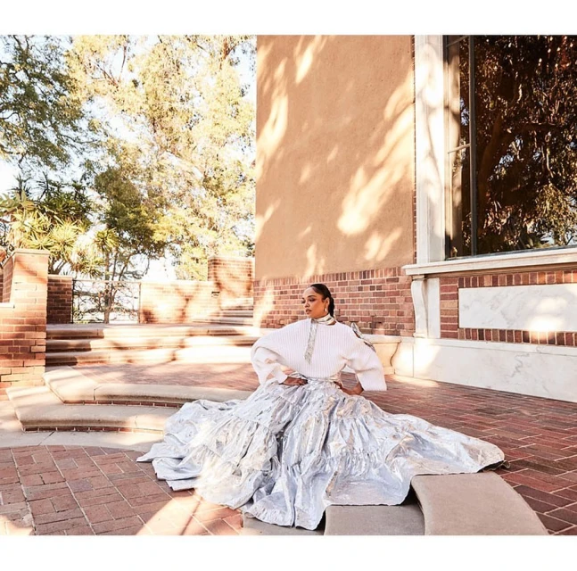 A woman poses in a white outfit with a silver skirt on a brick terrace, with buildings and trees in the background, under natural light.