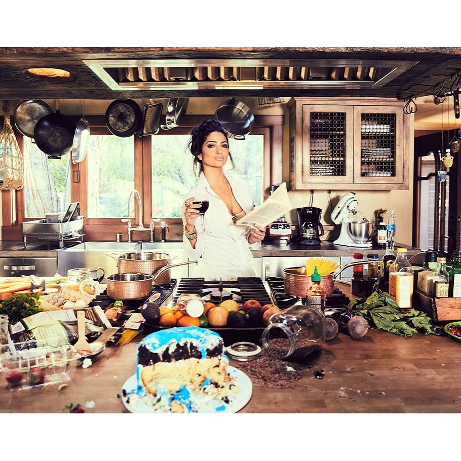 Woman in a white robe holds a wine glass and a book in a kitchen, surrounded by cooking ingredients, cookware, and a messy counter.
