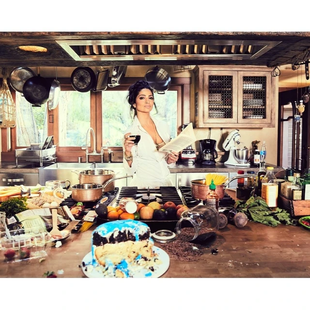 Woman in a white robe holds a wine glass and a book in a kitchen, surrounded by cooking ingredients, cookware, and a messy counter.