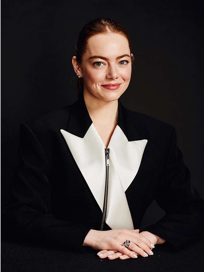 Close-up portrait of a woman with auburn hair, wearing a black and white tuxedo jacket, against a dark grey background.
