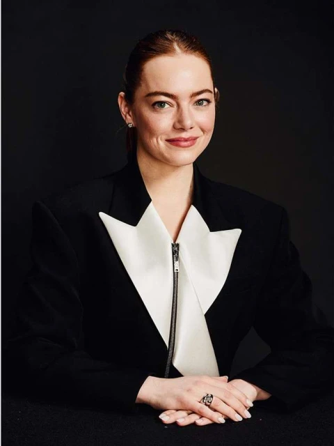 Close-up portrait of a woman with auburn hair, wearing a black and white tuxedo jacket, against a dark grey background.