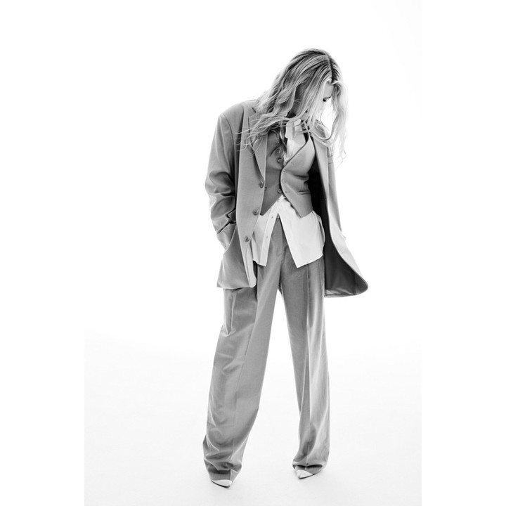Monochrome portrait of a model with long hair, wearing a three-piece suit, blouse, and pointed heels against a bright white backdrop.