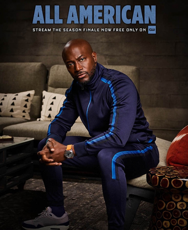 Promo poster of a bald adult male in a blue tracksuit sitting on a stool in a room with a couch and throw pillows.  'All American' text logo.