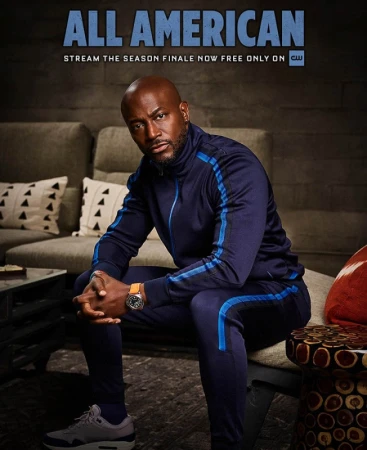 Promo poster of a bald adult male in a blue tracksuit sitting on a stool in a room with a couch and throw pillows.  'All American' text logo.