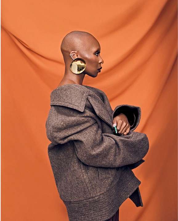 A bald African American woman in profile poses in a brown coat and gold earrings against a draped orange studio backdrop. Fashion portrait.