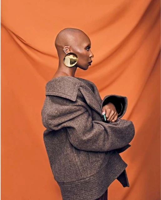 A bald African American woman in profile poses in a brown coat and gold earrings against a draped orange studio backdrop. Fashion portrait.