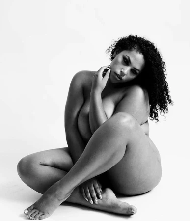Plus size woman with dark curly hair seated with legs crossed against a plain white background