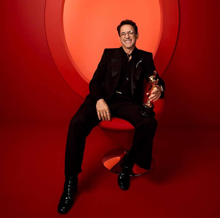 Robert Downey Jr. poses in a photo studio on a red chair holding his gold Oscar award. He is wearing a black suit and glasses.