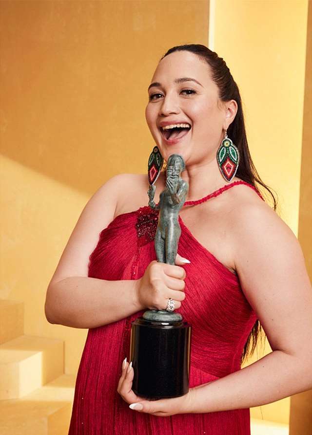 Adult female in a red dress holding an award and smiling in a photo studio setting with a tan background and medium-shot framing.