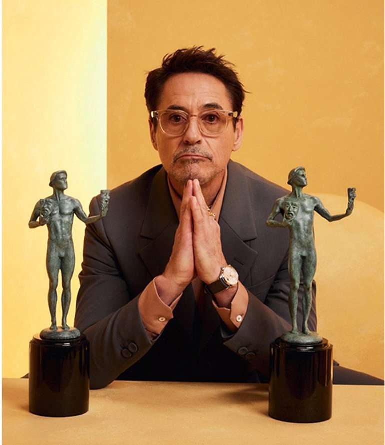 Robert Downey Jr. poses indoors with two SAG awards statues, wearing glasses and a grey suit with a gold-colored background.