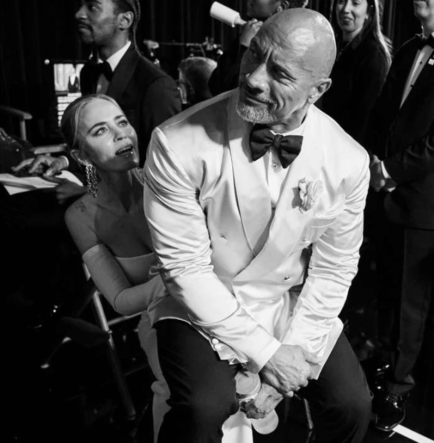 Black and white photo of Dwayne Johnson and Emily Blunt sitting and smiling during a backstage moment, likely at an awards show.