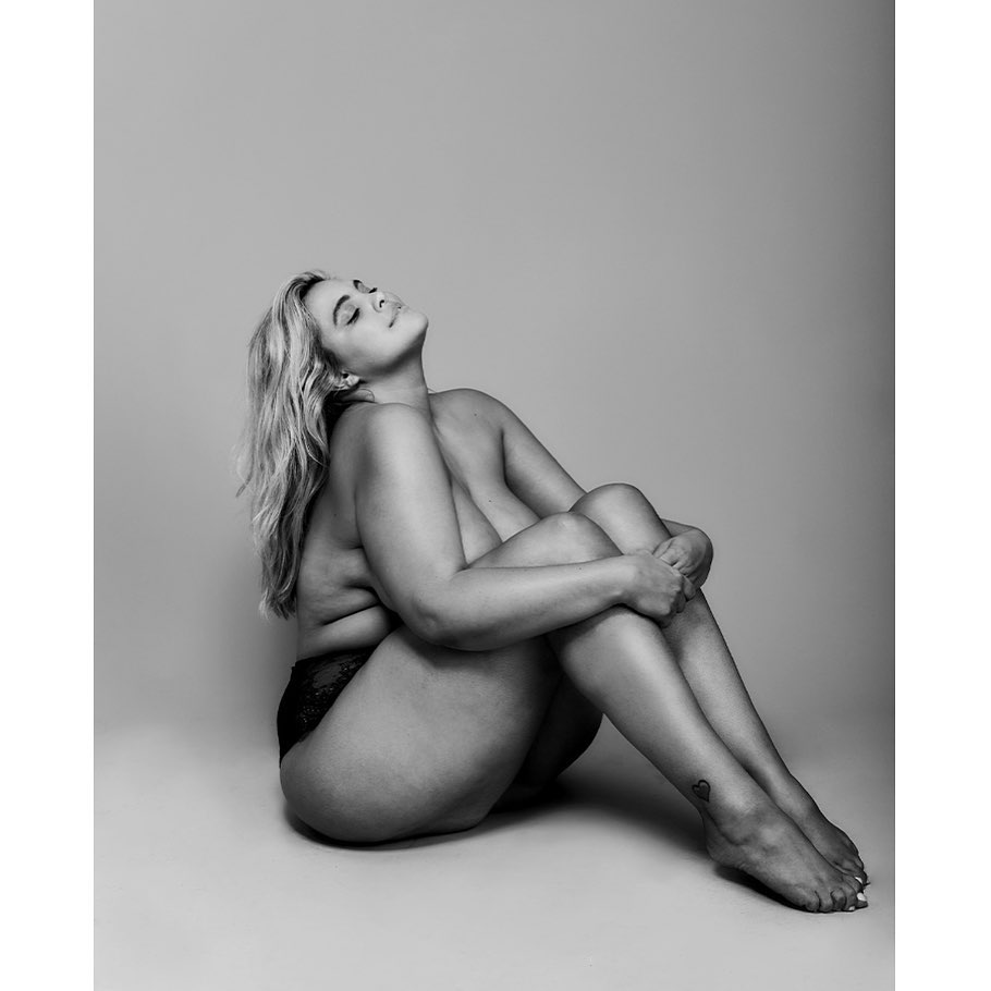 Monochrome studio shot of an adult female with blond hair, eyes closed, hugging her knees while sitting on the ground.