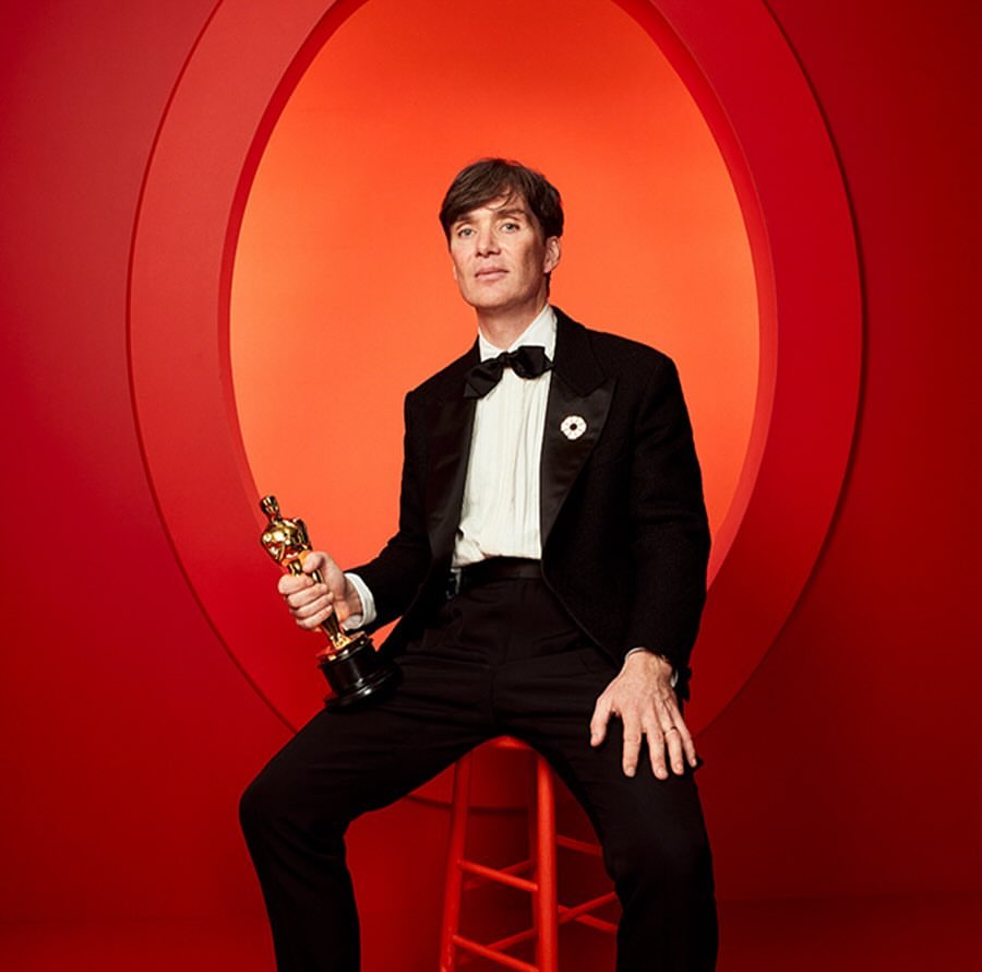 Cillian Murphy in a tuxedo holding an Oscar award, seated against a red and orange studio backdrop, captures an elegant moment.