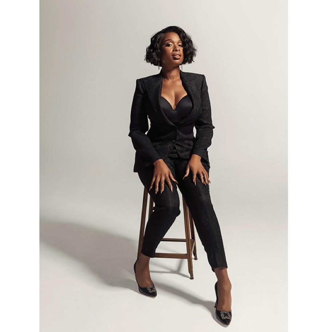 Jennifer Hudson sitting on a wooden stool wearing a black pantsuit and dark heels against a white backdrop.