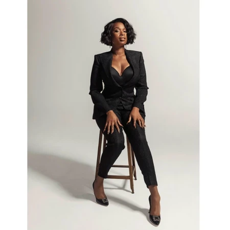 Jennifer Hudson sitting on a wooden stool wearing a black pantsuit and dark heels against a white backdrop.