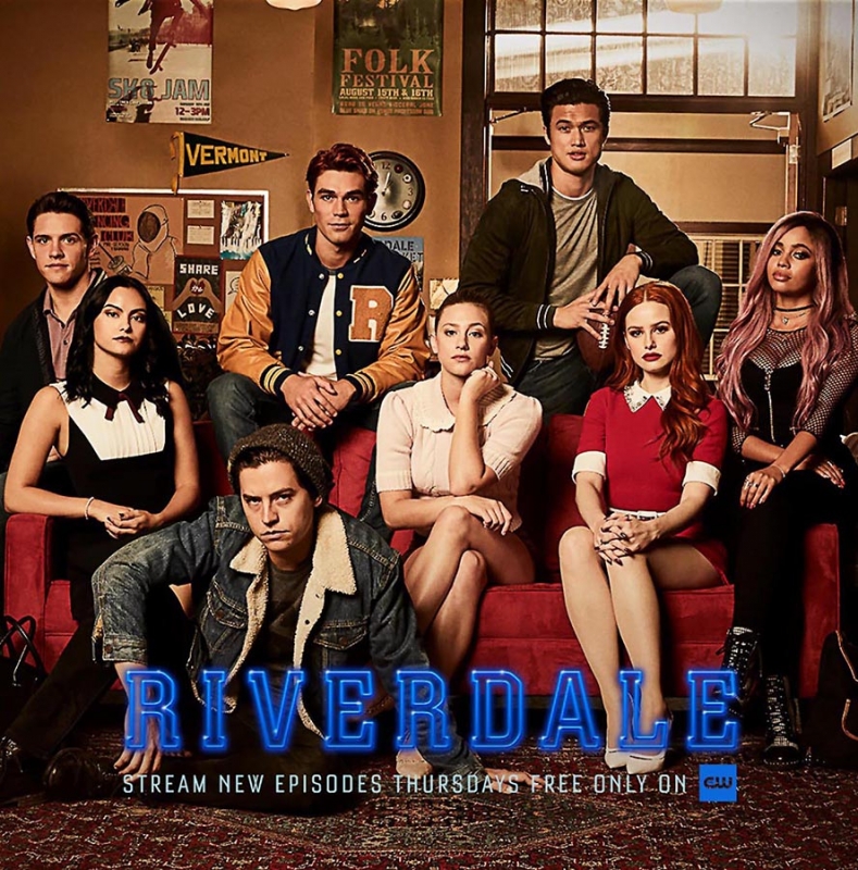 Group promotional image of the cast from the TV show Riverdale sitting on a red sofa inside with banners in the background.