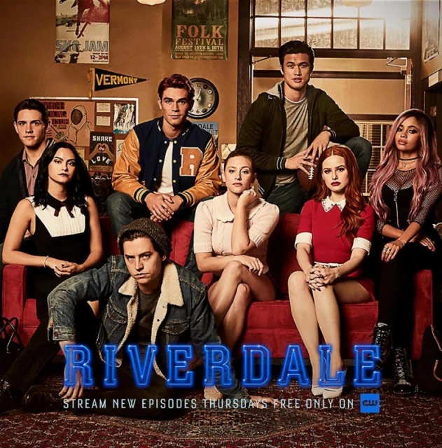 Group promotional image of the cast from the TV show Riverdale sitting on a red sofa inside with banners in the background.