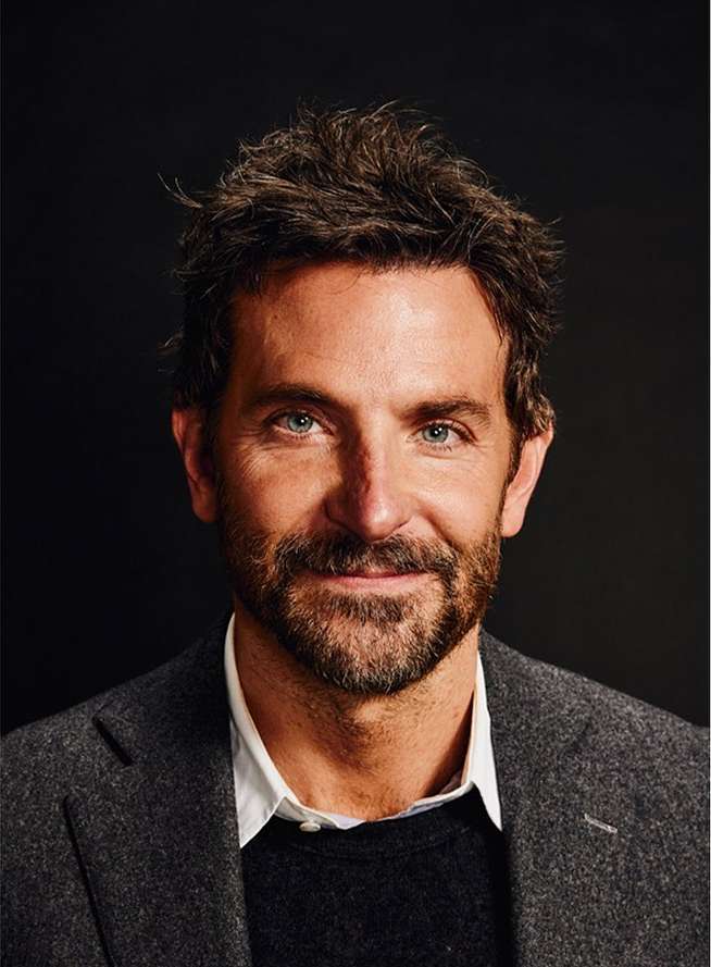 Headshot of an adult male with a beard, wearing a grey jacket and dark sweater, against a dark background.