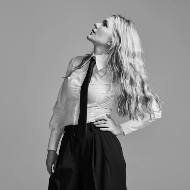 Black and white portrait of a woman in a white shirt, black tie, and wide-legged pants looking up, with long blonde wavy hair.