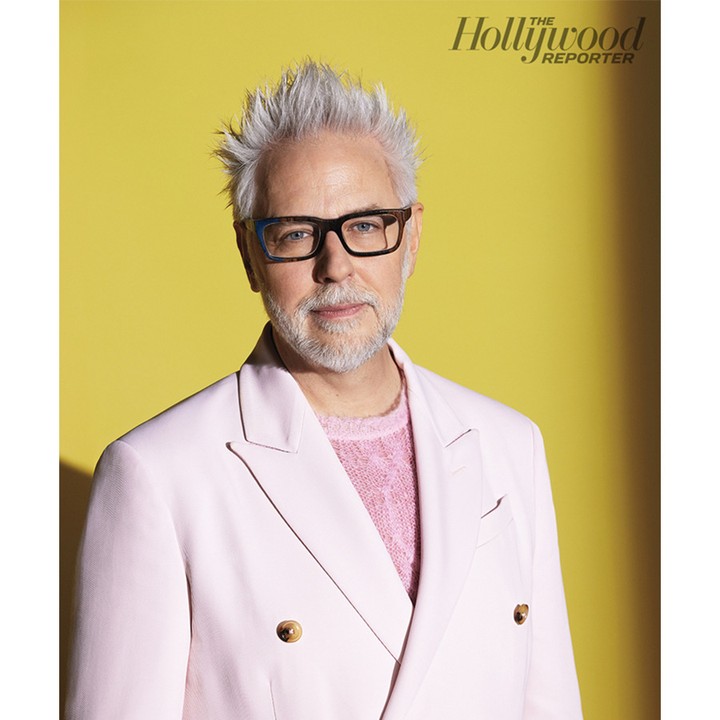 Headshot of James Gunn in pink blazer against yellow background. He wears glasses and has short gray hair in a studio setting.