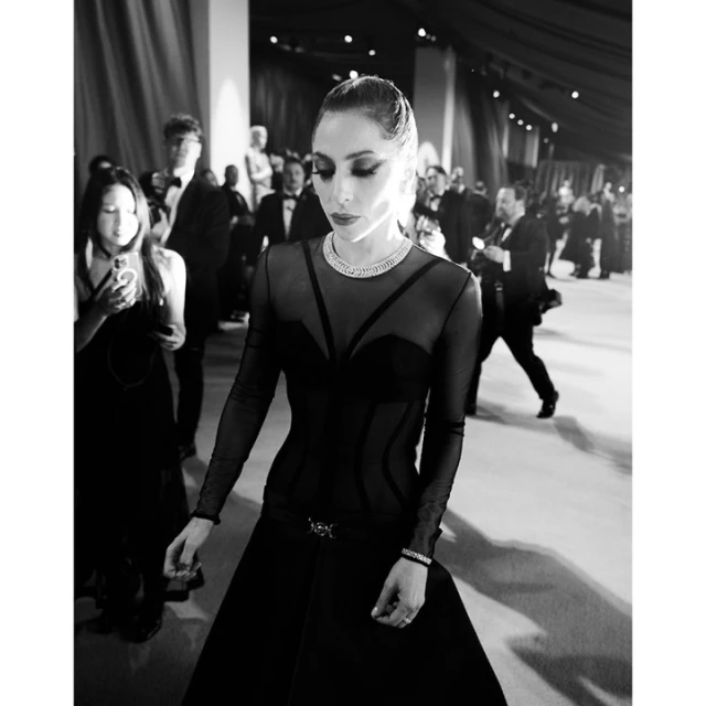A monochrome image captures a woman in a gown with sheer sleeves, standing on a carpeted surface surrounded by blurred figures in suits.