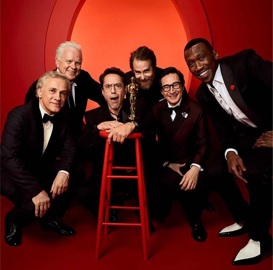 Group portrait of six men in tuxedos, one holding an Oscar award, gathered around a red stool against a red backdrop in a photo studio.