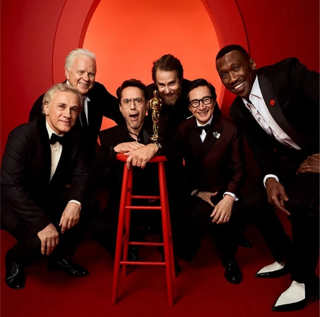 Group portrait of six men in tuxedos, one holding an Oscar award, gathered around a red stool against a red backdrop in a photo studio.