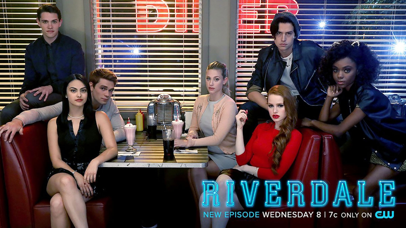 Group shot of Riverdale cast members sitting in diner booths, showcasing the show's stars in a retro-style setting with neon signage.