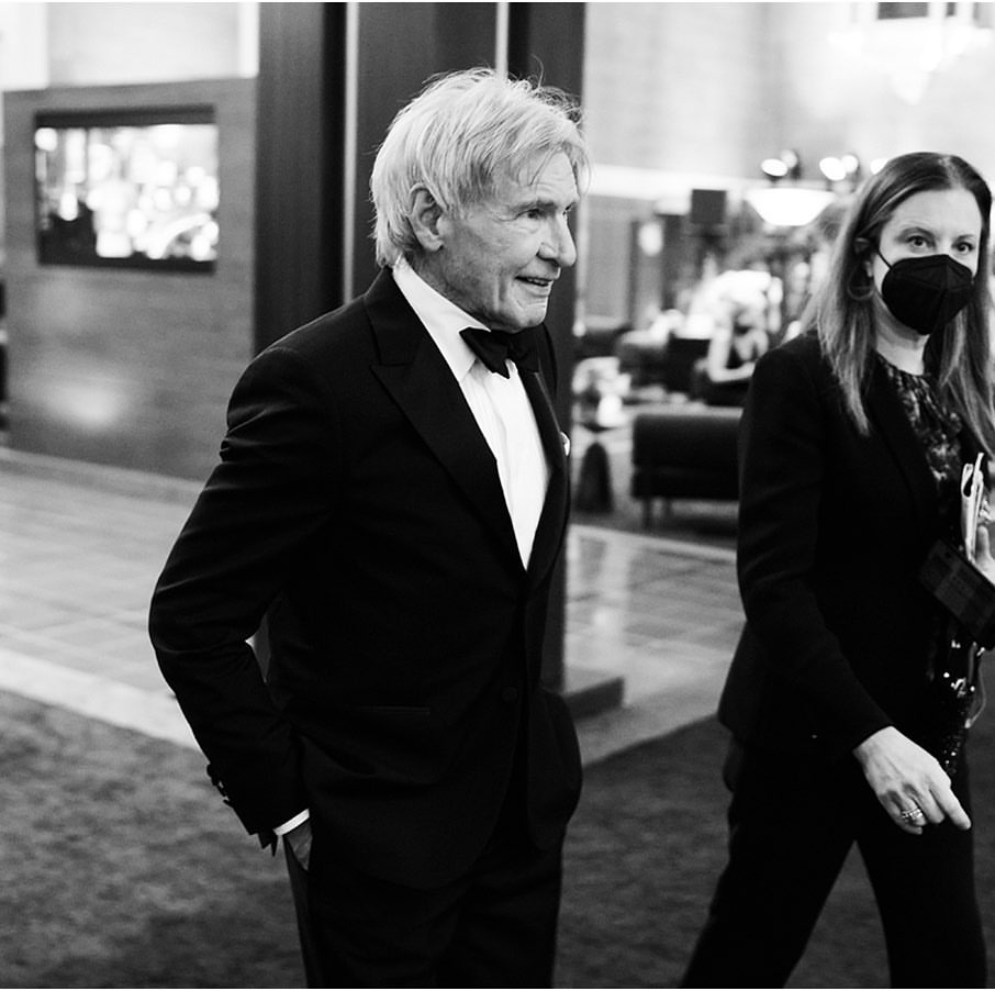 Black and white photo of an older adult male in a tuxedo with a woman in a mask standing in the background in a modern interior.