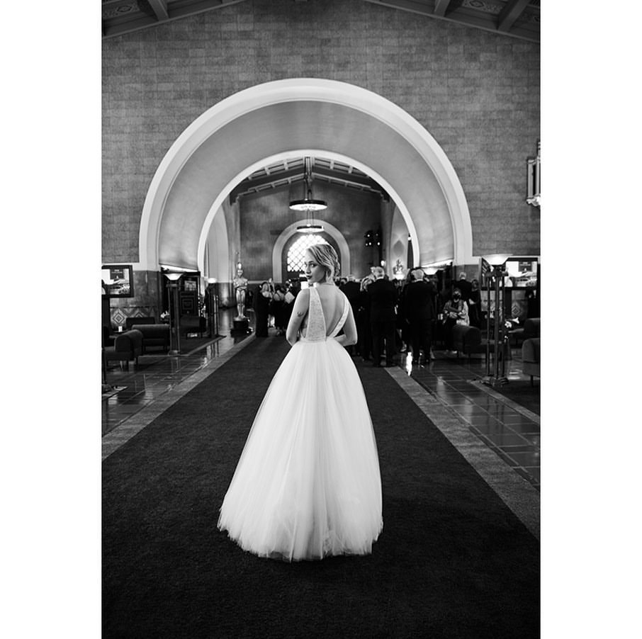 Black and white portrait of a bride in a wedding dress standing on a carpeted floor and looking back over her shoulder indoors.