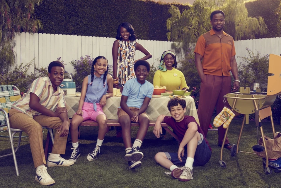 Outdoor portrait of a smiling family in retro clothing at a picnic table next to a grill, with green grass and a white fence.