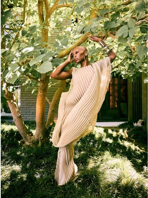 A woman poses outdoors wearing a beige pleated dress, surrounded by greenery and trees in a fashion shot.