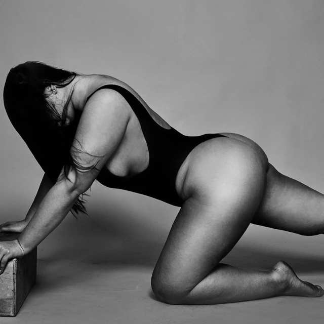 Monochrome studio shot of a woman in a black one-piece swimsuit, kneeling on a wooden cube, showcasing a sensual pose and figure.