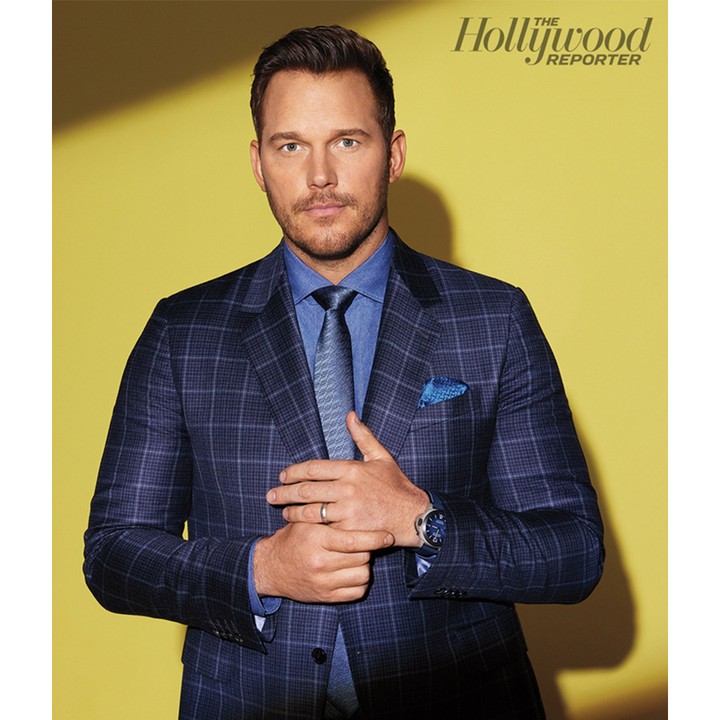 Actor Chris Pratt poses in a blue plaid suit, light blue shirt and patterned tie in a studio portrait against a yellow background.