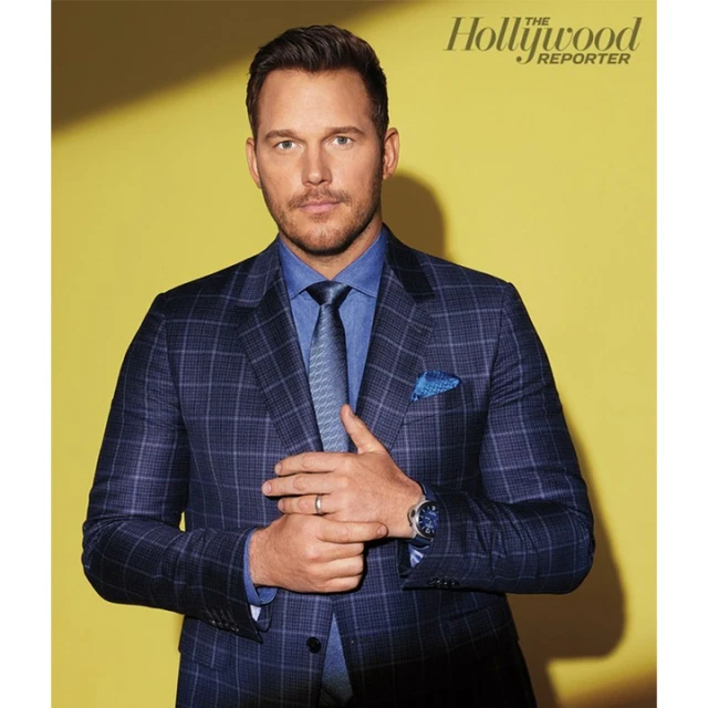 Actor Chris Pratt poses in a blue plaid suit, light blue shirt and patterned tie in a studio portrait against a yellow background.