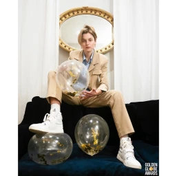 Portrait of Emma Corrin sitting on a dark sofa holding a clear balloon filled with gold confetti, photographed by Matt Sayles.