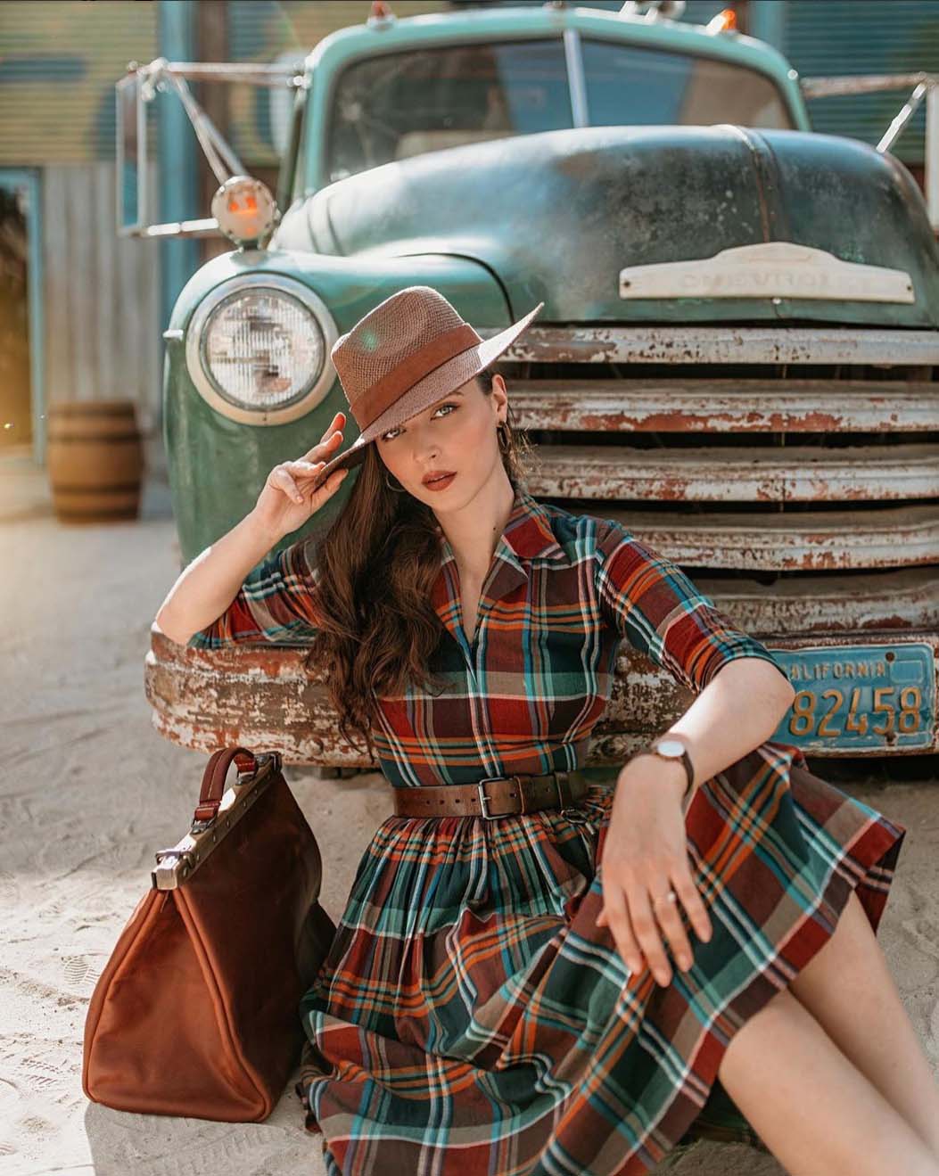 Young woman in plaid dress poses by a vintage truck in an outdoor setting with a brown handbag, hat, and watch.