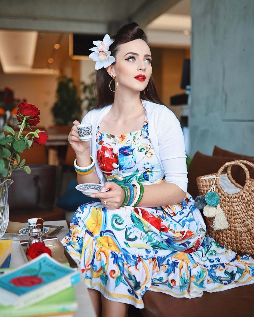 A young woman with retro hair and makeup, wearing a floral dress, sits indoors holding a teacup near a table with books and roses.