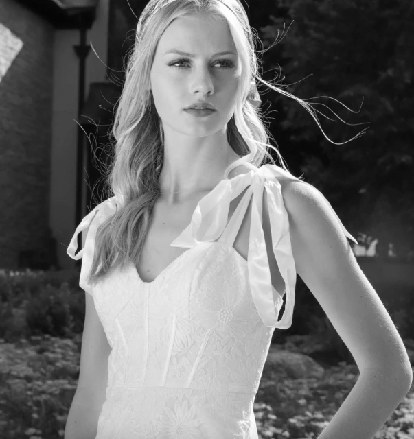 Monochrome portrait of a blonde woman wearing a white dress with ribbon straps, set outdoors against a blurred background of foliage.