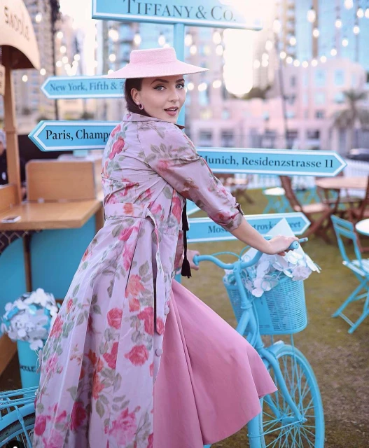Adult female rides a light blue bicycle while posing near a Tiffany & Co. sign with the names of international cities, outside on grass.
