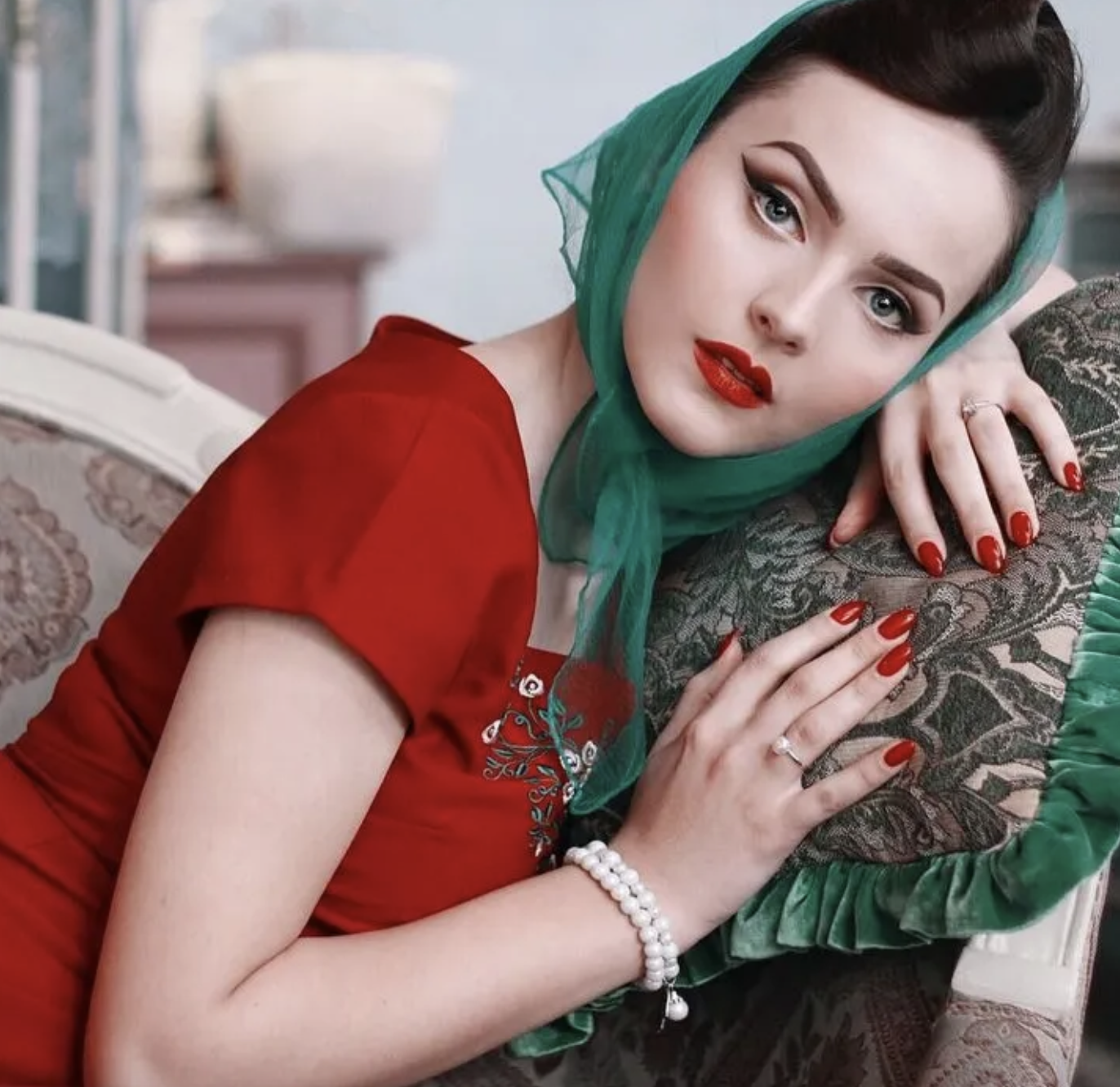 A young woman with fair skin poses indoors, wearing a red dress and green headscarf, leaning against a decorative pillow with bright red lips.